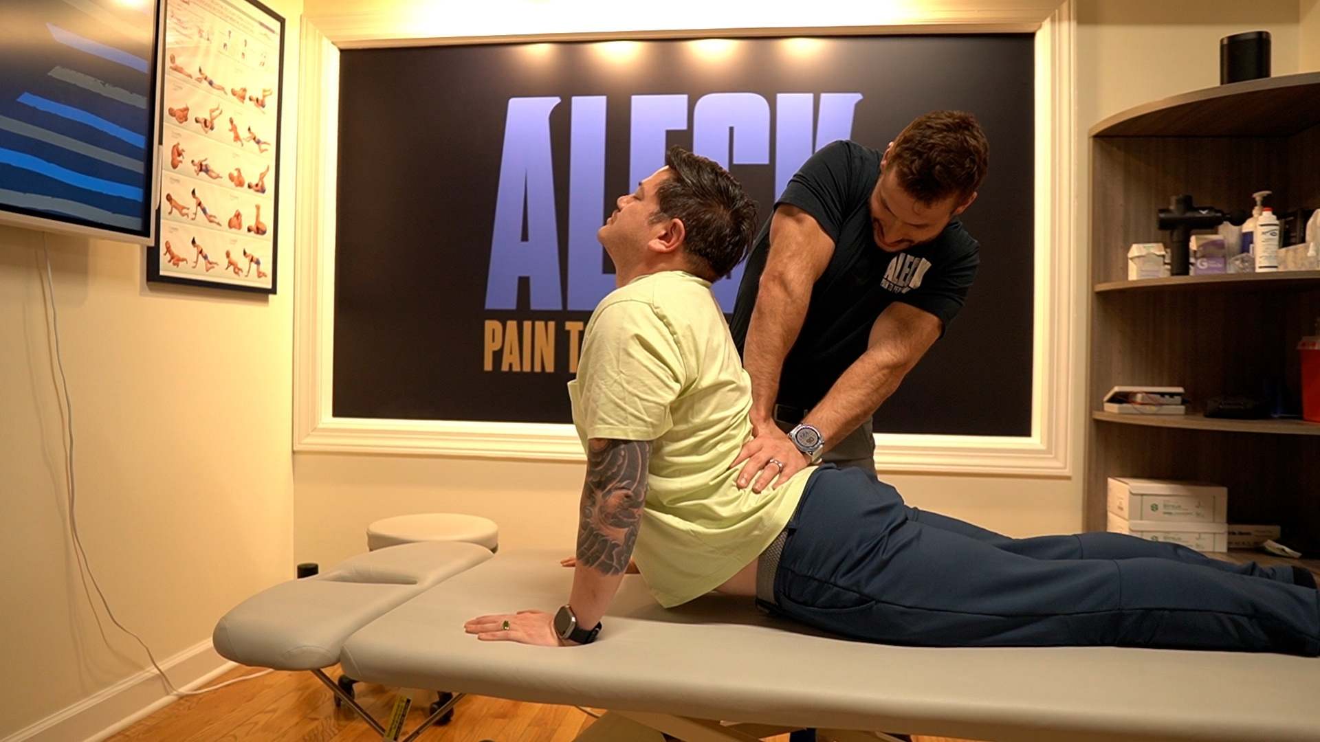 A chiropractor adjusts the lower back of a patient performing a cobra stretch on a light gray examination table in a professional clinic. The setting includes a large