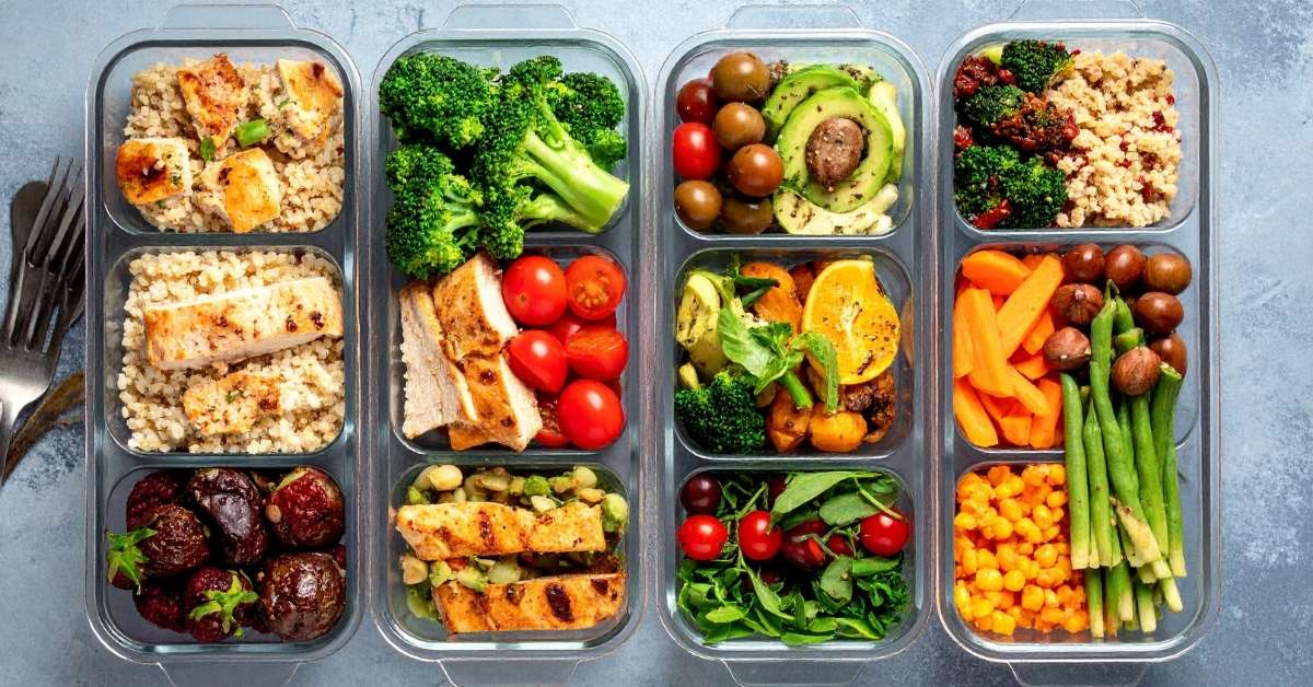 Overhead view of four glass meal prep containers filled with grilled chicken, grains, vegetables, and salad ingredients arranged on a light surface.