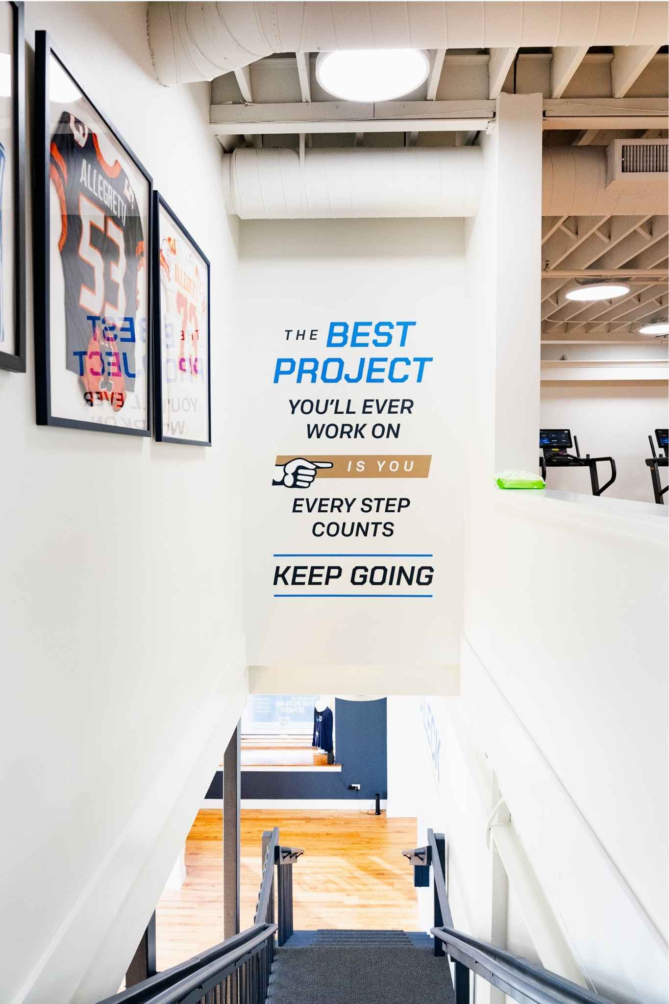 Modern gym stairwell featuring a large motivational wall mural that reads “The best project you’ll ever work on is you. Every step counts. Keep going,” with framed football jerseys displayed along the left wall.